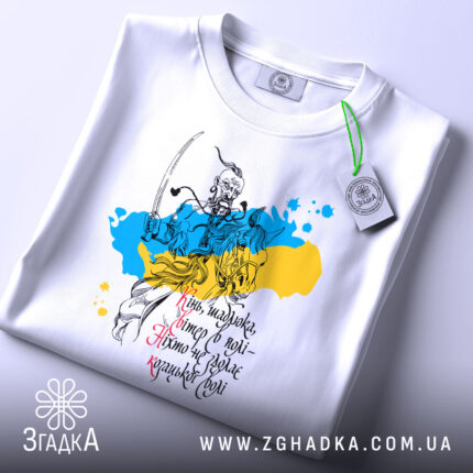 Футболка козацька воля, біла, складена на столі, фон світло-сірий, принт з козацьким воїном на фоні жовто-блакитних плям, текст українською мовою, з зеленим ярликом.