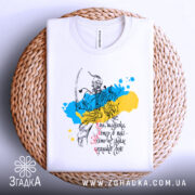 Футболка козацька воля, біла, складена на плетеній кошику, фон світло-білий, принт з козацьким воїном на фоні жовто-блакитних плям, текст українською мовою.