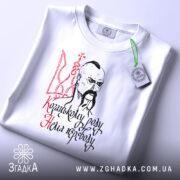 Футболка козацькому роду нема переводу. Біла футболка, складена на поверхні, зображена з верхнього ракурсу. На футболці видно принт з козацьким малюнком та написом, а також зелену етикетку.