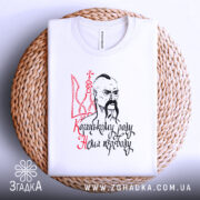 Футболка козацькому роду нема переводу. Біла футболка, складена на плетеній основі, зображена з верхнього ракурсу. Принт з козацьким малюнком та написом добре видно на фоні.