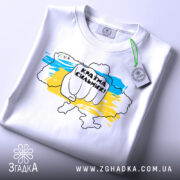 Футболка країна незламних, біла, складена на столі, фон світлий, принт з рукою, що тримає напис "Країна сильних!" на фоні карти України в жовто-блакитних кольорах, з етикеткою на комірці.