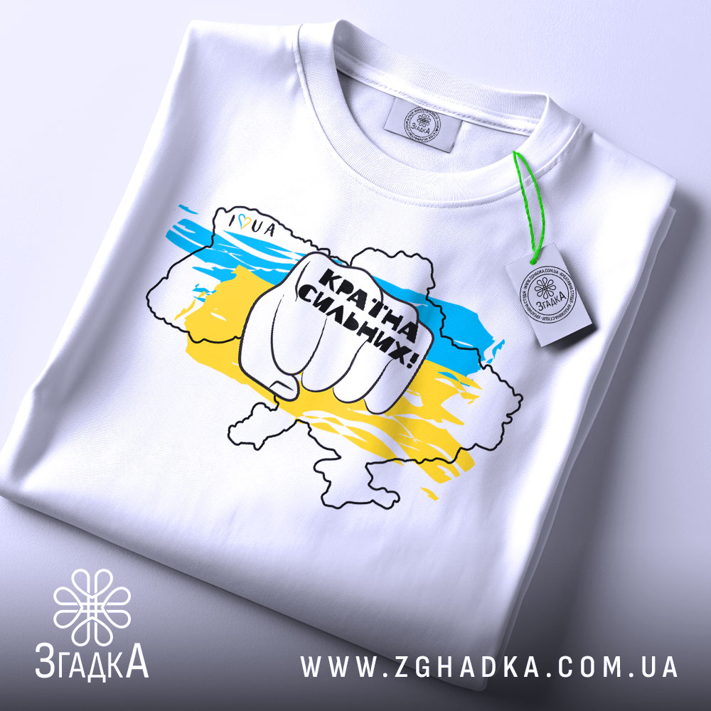 Футболка країна незламних, біла, складена на столі, фон світлий, принт з рукою, що тримає напис "Країна сильних!" на фоні карти України в жовто-блакитних кольорах, з етикеткою на комірці.