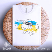 Футболка країна незламних, біла, складена на плетеній кошику, фон світлий, принт з рукою, що тримає напис "Країна сильних!" на фоні карти України в жовто-блакитних кольорах.