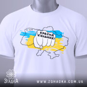 Футболка країна незламних, біла, складена на столі, фон світлий, принт з рукою, що тримає напис "Країна сильних!" на фоні карти України в жовто-блакитних кольорах, з етикеткою на комірці.