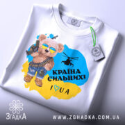 Футболка країна вільних, біла, зображена на столі, фон світло-сірий, принт з ведмедиком у сонцезахисних окулярах, з автоматом і гелікоптером, текст "Країна сильних! I ♥ UA" у жовто-блакитному кольорі, з етикеткою на комірці.