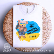 Футболка країна вільних, біла, зображена на плетеній кошику, фон світло-сірий, принт з ведмедиком у сонцезахисних окулярах, з автоматом і гелікоптером, текст "Країна сильних! I ♥ UA" у жовто-блакитному кольорі.