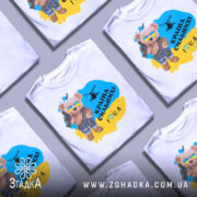 Футболка країна вільних, біла, зображена на манекені, фон світло-сірий, принт з ведмедиком у сонцезахисних окулярах, з автоматом і гелікоптером, текст "Країна сильних! I ♥ UA" у жовто-блакитному кольорі.