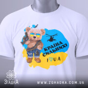 Футболка країна вільних, біла, зображена на столі, фон світло-сірий, принт з ведмедиком у сонцезахисних окулярах, з автоматом і гелікоптером, текст "Країна сильних! I ♥ UA" у жовто-блакитному кольорі, з етикеткою на комірці.