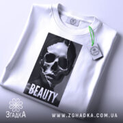Футболка краса без ілюзій, білого кольору, складена на поверхні, фон світло-сірий, принт з черепом та написом 'BEAUTY' розташований по центру, з етикеткою, що звисає з коміра.