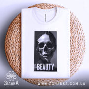Футболка краса без ілюзій, білого кольору, складена на плетеній кошику, фон світло-сірий, принт з черепом та написом 'BEAUTY' розташований по центру, виглядає елегантно.