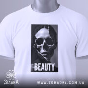 Футболка краса без ілюзій, білого кольору, складена на поверхні, фон світло-сірий, принт з черепом та написом 'BEAUTY' розташований по центру, з етикеткою, що звисає з коміра.
