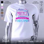 Футболка кращі матусі, білого кольору, зображена з фронтального ракурсу на манекені, фон нейтральний, принт містить текст у яскравих кольорах: фіолетовий, рожевий та блакитний, оформлений декоративними елементами.