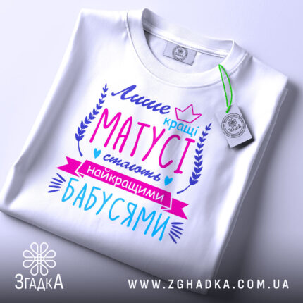 Футболка кращі матусі, білого кольору, зображена зверху на плоскій поверхні, фон світлий, принт яскравий з текстом у фіолетових, рожевих та блакитних відтінках, з етикеткою на рукаві.