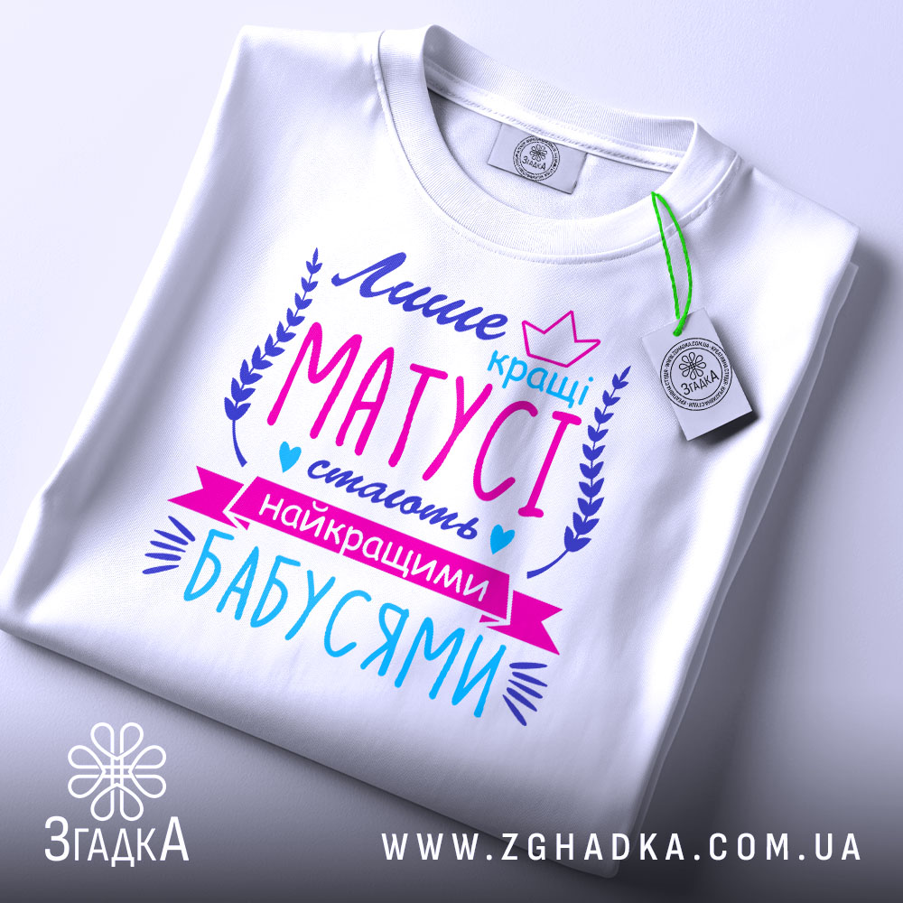 Футболка кращі матусі, білого кольору, зображена зверху на плоскій поверхні, фон світлий, принт яскравий з текстом у фіолетових, рожевих та блакитних відтінках, з етикеткою на рукаві.