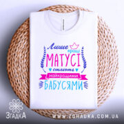 Футболка кращі матусі, білого кольору, розкладена на плоскій поверхні з плетеним елементом на фоні, принт яскравий з текстом у фіолетових, рожевих та блакитних кольорах, оформлений декоративними елементами.
