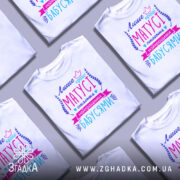 Футболка кращі матусі, білого кольору, зображена з фронтального ракурсу на манекені, фон нейтральний, принт містить текст у яскравих кольорах: фіолетовий, рожевий та блакитний, оформлений декоративними елементами.