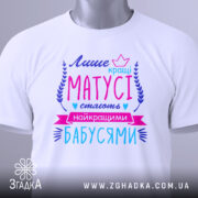 Футболка кращі матусі, білого кольору, зображена зверху на плоскій поверхні, фон світлий, принт яскравий з текстом у фіолетових, рожевих та блакитних відтінках, з етикеткою на рукаві.