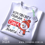 Футболка кращого сина. Біла футболка, складена на столі, з яскравим принтом сніговика в червоній шапці. Текст "Хочу півсвіту обійди, кращого сина не знайти" розташований на футболці. На футболці є етикетка з логотипом бренду. Фон світлий, що підкреслює деталі принта.