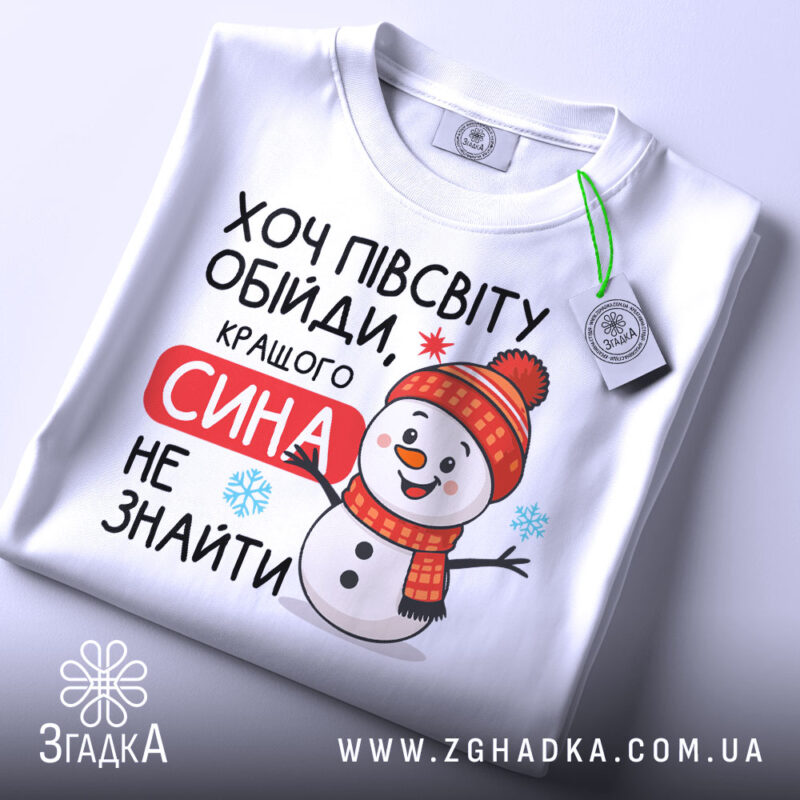 Футболка кращого сина. Біла футболка, складена на столі, з яскравим принтом сніговика в червоній шапці. Текст "Хочу півсвіту обійди, кращого сина не знайти" розташований на футболці. На футболці є етикетка з логотипом бренду. Фон світлий, що підкреслює деталі принта.