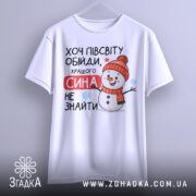 Футболка кращого сина. Біла футболка, підвішена на вішалці, з принтом сніговика у червоній шапці. Текст "Хочу півсвіту обійди, кращого сина не знайти" яскраво виділяється на фоні. Фон світло-сірий, що робить принт більш помітним.