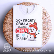 Футболка кращого сина. Біла футболка, складена на плетеній кошику, з принтом сніговика в червоній шапці. Текст "Хочу півсвіту обійди, кращого сина не знайти" розташований на футболці. Фон нейтральний, що підкреслює текст та малюнок.