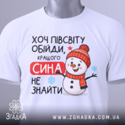 Футболка кращого сина. Біла футболка, складена на столі, з яскравим принтом сніговика в червоній шапці. Текст "Хочу півсвіту обійди, кращого сина не знайти" розташований на футболці. На футболці є етикетка з логотипом бренду. Фон світлий, що підкреслює деталі принта.