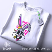 Футболка крейзі заєць, біла, лежить на столі, фон світло-сірий, принт з зайцем з короною, яскраві кольори, на футболці видно етикетку з зеленим шнурком.