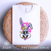 Футболка крейзі заєць, біла, лежить на плетеній кошику, фон світло-сірий, принт з зайцем з короною, яскраві кольори, деталі принта включають рожеві та жовті елементи.