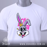 Футболка крейзі заєць, біла, лежить на столі, фон світло-сірий, принт з зайцем з короною, яскраві кольори, на футболці видно етикетку з зеленим шнурком.