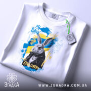 Футболка кролик будь собою. Біла футболка, зображена зверху, з принтом кролика в чорній куртці на жовто-блакитному фоні. На футболці є етикетка з зеленим шнурком.