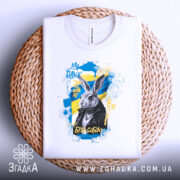 Футболка кролик будь собою. Біла футболка, складена на плетеній основі, з принтом кролика в чорній куртці на жовто-блакитному фоні. Текст "БУДЬ СОБОЮ" та інші елементи принта добре помітні.