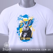 Футболка кролик будь собою. Біла футболка, зображена зверху, з принтом кролика в чорній куртці на жовто-блакитному фоні. На футболці є етикетка з зеленим шнурком.