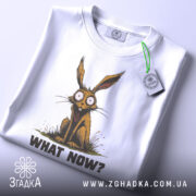 Футболка кролик карикатура, біла, зображена з верхнього ракурсу, на світлому фоні, принт з кроликом, що сидить, з текстом 'WHAT NOW?' та етикеткою на рукаві.
