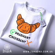 Футболка круасан стиль, біла, складена на столі, фон світло-сірий, принт з оранжевим круасаном та текстом 'PRASHANT CROISSANT' чорного кольору з зеленим галочкою, на футболці є етикетка.