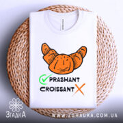 Футболка круасан стиль, біла, складена на плетеному кошику, фон світло-сірий, принт з оранжевим круасаном та текстом 'PRASHANT CROISSANT' чорного кольору з зеленим галочкою.