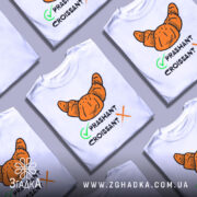 Футболка круасан стиль, біла, зображена на манекені, фон світло-сірий, принт з оранжевим круасаном та текстом 'PRASHANT CROISSANT' чорного кольору з зеленим галочкою.