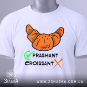 Футболка круасан стиль, біла, складена на столі, фон світло-сірий, принт з оранжевим круасаном та текстом 'PRASHANT CROISSANT' чорного кольору з зеленим галочкою, на футболці є етикетка.