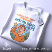 Футболка кругом якась фігня, біла футболка складена на столі, ракурс зверху, фон світлий, принт з зображенням чоловіка з рудим волоссям, який прикриває очі від сонця, текст зверху оранжевого кольору, текст внизу блакитного кольору, на футболці зелена етикетка.