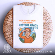 Футболка кругом якась фігня, біла футболка складена на плетеній основі, ракурс зверху, фон нейтральний, принт з зображенням чоловіка з рудим волоссям, який прикриває очі від сонця, текст зверху оранжевого кольору, текст внизу блакитного кольору.