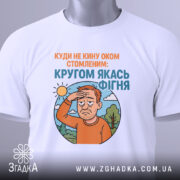 Футболка кругом якась фігня, біла футболка складена на столі, ракурс зверху, фон світлий, принт з зображенням чоловіка з рудим волоссям, який прикриває очі від сонця, текст зверху оранжевого кольору, текст внизу блакитного кольору, на футболці зелена етикетка.