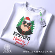 Футболка крутого Нового Року, біла, зображена зверху, фон світлий, принт з персонажем у червоній шапці та ялинками, текст 'Крутого Нового року!' яскравими кольорами, на футболці є етикетка.