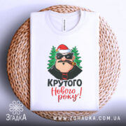 Футболка крутого Нового Року, біла, зображена зверху на плетеній основі, фон світлий, принт з персонажем у червоній шапці та ялинками, текст 'Крутого Нового року!' яскравими кольорами.