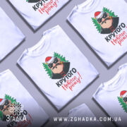 Футболка крутого Нового Року, біла, зображена з близької відстані, фон нейтральний, принт з веселим персонажем у червоній шапці та ялинками, текст 'Крутого Нового року!' яскравими кольорами.
