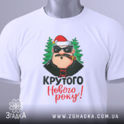 Футболка крутого Нового Року, біла, зображена зверху, фон світлий, принт з персонажем у червоній шапці та ялинками, текст 'Крутого Нового року!' яскравими кольорами, на футболці є етикетка.