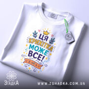 Футболка крихітка може все. Складена біла футболка з кольоровим принтом, розташованим на верхній частині, на фоні світло-сірого кольору. Принт містить текст "ЦЯ КРИХІТКА МОЖЕ ВСЕ!" та прикрашений елементами, такими як корона і листя, з етикеткою, що звисає з коміра.