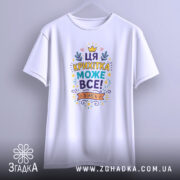 Футболка крихітка може все. Біла футболка з кольоровим принтом, що висить на вішалці, на фоні світло-сірого кольору. Принт містить текст "ЦЯ КРИХІТКА МОЖЕ ВСЕ!" з яскравими елементами, такими як зірочки та листя.