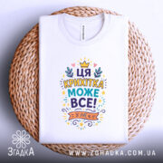 Футболка крихітка може все. Складена біла футболка з кольоровим принтом, розташованим на верхній частині, на фоні плетеного кошика. Принт містить текст "ЦЯ КРИХІТКА МОЖЕ ВСЕ!" оточений кольоровими елементами, такими як корона і листя.