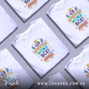 Футболка крихітка може все. Біла футболка з кольоровим принтом, зображеним на грудях, на фоні світло-сірого градієнту. Принт містить текст "ЦЯ КРИХІТКА МОЖЕ ВСЕ!" оточений кольоровими елементами, такими як зірочки та листя.