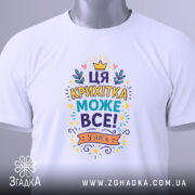 Футболка крихітка може все. Складена біла футболка з кольоровим принтом, розташованим на верхній частині, на фоні світло-сірого кольору. Принт містить текст "ЦЯ КРИХІТКА МОЖЕ ВСЕ!" та прикрашений елементами, такими як корона і листя, з етикеткою, що звисає з коміра.