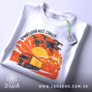 Футболка кримський міст горить, біла, складена на поверхні, фон нейтральний, принт зображає вантажівку та дрони на фоні палаючого мосту з оранжевими та червоними відтінками, з етикеткою на рукаві.